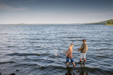 Fototapeta na wymiar Young man and teenage boy standing in the lake and throwing stones into water on summer day