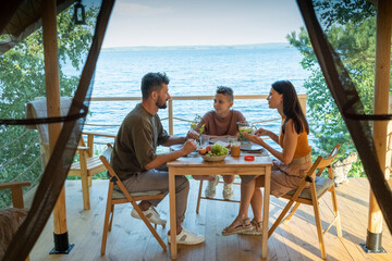 Happy family of three having dinner by wooden table on terrace by seaside