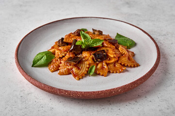Pasta farfalle with eggplant, tomato sauce, cheese and basil on plate on a gray textured background, close up