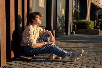 Fototapeta premium Teenage skateboarder sitting by wall on walkside and having rest
