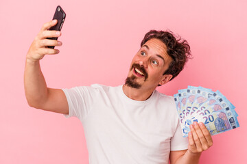 Young caucasian man holding bills and piggy bank isolated on pink background