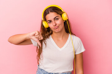 Young caucasian woman listening to music isolated on pink background  showing a dislike gesture,...