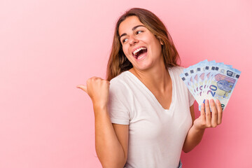 Young caucasian woman holding bills isolated on pink background  points with thumb finger away, laughing and carefree.