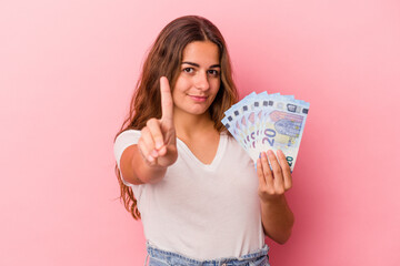 Young caucasian woman holding bills isolated on pink background  showing number one with finger.