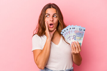 Young caucasian woman holding bills isolated on pink background  is saying a secret hot braking news and looking aside