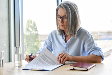 Serious older lawyer businesswoman in glasses reading signing trust partnership contract sit at...