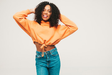Beautiful black woman with afro curls hairstyle.Smiling model in orange hoodie and trendy jeans clothes. Sexy carefree female posing on white background in studio. Tanned and cheerful. Winks