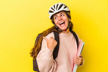 Young caucasian student woman wearing a bike helmet isolated on yellow background  points with thumb finger away, laughing and carefree.