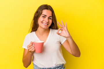 Young caucasian woman holding a mug isolated on yellow background  cheerful and confident showing ok gesture.