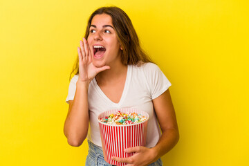 Young caucasian woman holding a popcorns isolated on yellow background  shouting and holding palm near opened mouth.