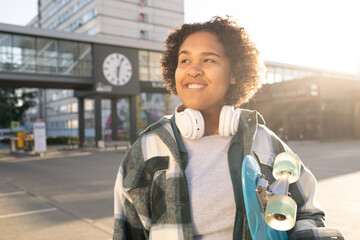 Cute cheerful teenager with skateboard and headphones standing in urban environment