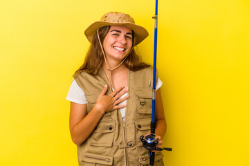 Young caucasian fisherwoman holding a rod isolated on yellow background  laughs out loudly keeping hand on chest.