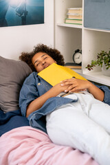 Tired teenage girl with open book on her chest sleeping by shelf in bedroom