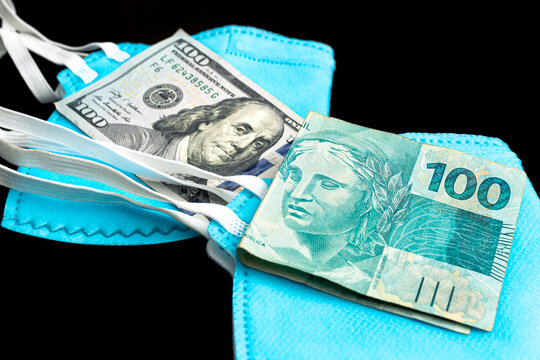 Cédula do Real Brasileiro de 100 Reais e uma Cédula de 100 Dólares dos Estados Unidos que estão sobre máscaras n95 com fundo preto. Crise econômica no Brasil durante a pandemia do novo coronavírus.