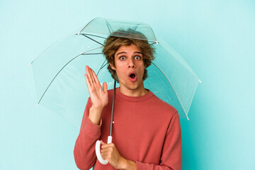 Young caucasian man with makeup holding umbrella isolated on blue background  surprised and shocked.
