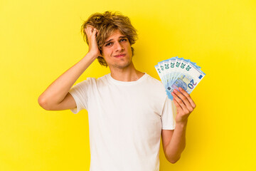 Young caucasian man with makeup holding bills isolated on yellow background  being shocked, she has remembered important meeting.