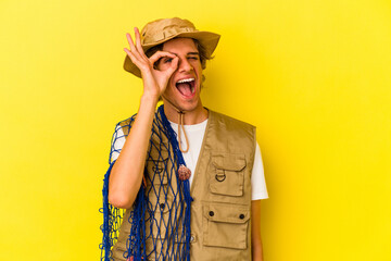Young fisherman with makeup holding a net isolated on yellow background  excited keeping ok gesture on eye.
