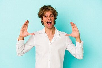 Young caucasian man with make up isolated on blue background  holding something with palms, offering to camera.