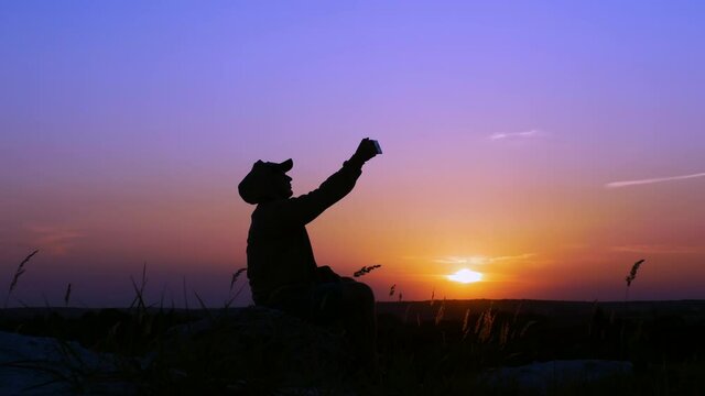 A lonely man on the top of a mountain taking a selfie, a teenager taking pictures of himself at sunset. Silhouette of a man at an orange sunset.