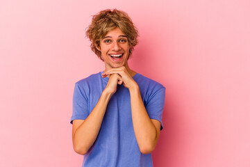 Young caucasian man with make up isolated on pink background keeps hands under chin, is looking happily aside.