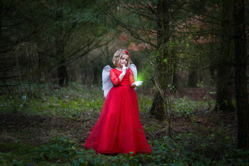 Fototapeta premium Kleines Mädchen, kleiner Engel steht im Wald mit ihrem roten Kleid, weißen Flügeln. Sie hält Zauber grünes Licht in der Hand, es leuchtet, Sie hält ihren Zeigefinger vor Mund und bietet leiser zu sein
