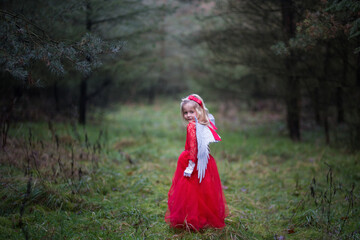 Kleines Mädchen, kleiner Engel steht im Wald mit ihrem roten Kleid, weißen Flügeln, rote Schleife in dem Haar,  sieht hübsch aus und hat blonde Haare. Sie steht mit dem rücken zu uns, dreht sich um.