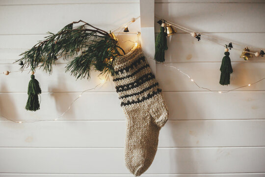 Christmas Rustic Stocking With Fir Branch And Stylish Garland With Bells And Trees On White Wooden Wall With Christmas Lights. Festive Decorated Atmospheric Scandinavian Room. Modern Handmade Decor