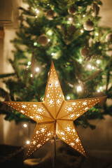 Christmas star on background of christmas tree in lights in evening room. Big paper star garland glowing in festive decorated scandinavian room. Atmospheric christmas eve. Happy holidays