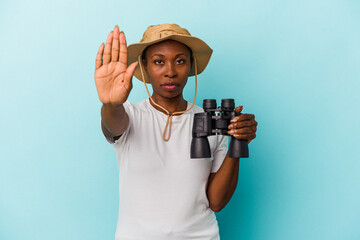 Young african american woman holding binoculars isolated on blue background standing with outstretched hand showing stop sign, preventing you.