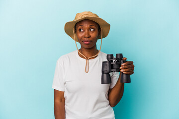 Young african american woman holding binoculars isolated on blue background confused, feels doubtful and unsure.