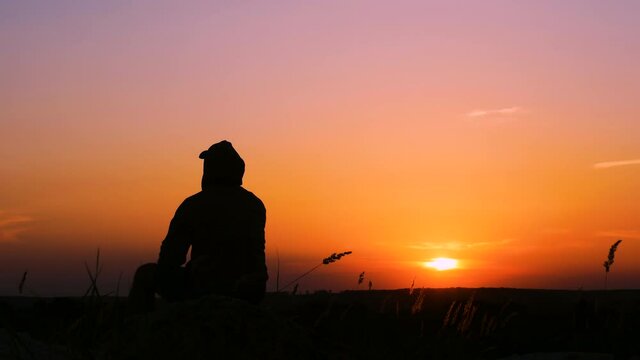 Sad silhouette of a man on a mountain at an orange sunrise or sunset. A sad or pensive person on a mountain, a depressed state of a person at sunset or daydreaming.