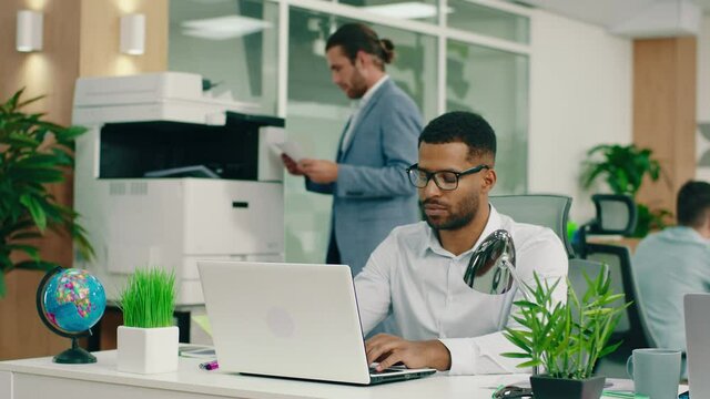 A very alluring manager is sitting at his desk in the big office wearing black glasses as he does his work on the laptop another man in the back is at the printer