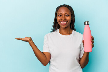 Young african american woman holding canteen isolated on blue background showing a copy space on a palm and holding another hand on waist.