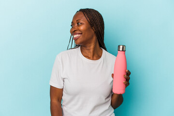 Young african american woman holding canteen isolated on blue background looks aside smiling, cheerful and pleasant.