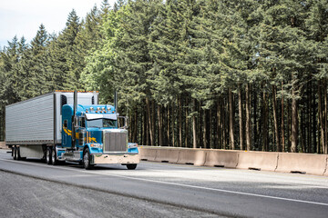 Classic stylish blue big rig semi truck with lot of chrome details transporting cargo in reefer semi trailer driving on the highway road with green forest on the side