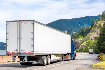 Convoy of the big rigs semi trucks with dry van semi trailers transporting cargo driving on the winding road with river and mountain on the sides © vit