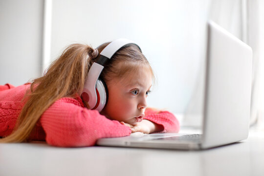 Cute little girl in headphones seriously using laptop at home for online education, home studying and online communication. Kids distance learning. Social distance. Stay at home.
