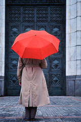 A woman under a red umbrella stands alone at the door of an old house