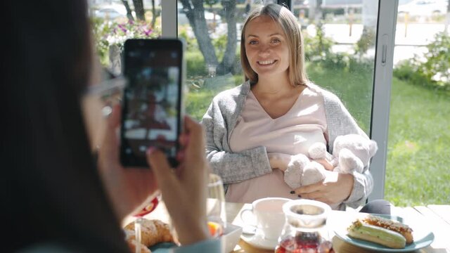 Happy pregnant woman is posing for smartphone camera with teddy bear while friends are taking pictures in cafe. Pregnancy and photography concept.