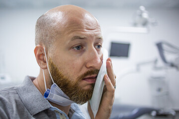 Unhappy and scared caucasian male in a dental clinic waiting for a check up and holding a cooling element at his cheek because of a toothache