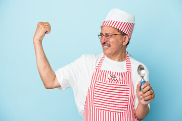 Senior indian ice cream man holding a scoop isolated on blue background raising fist after a victory, winner concept.