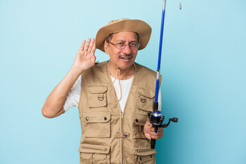 Senior indian fisherman holding rod isolated on blue background trying to listening a gossip.