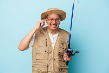 Senior indian fisherman holding rod isolated on blue background covering ears with hands.