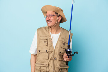 Senior indian fisherman holding rod isolated on blue background looks aside smiling, cheerful and pleasant.