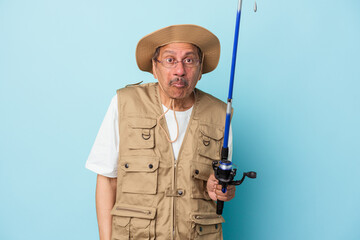 Senior indian fisherman holding rod isolated on blue background shrugs shoulders and open eyes confused.