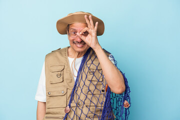 Senior indian fisherman holding net isolated on blue background excited keeping ok gesture on eye.