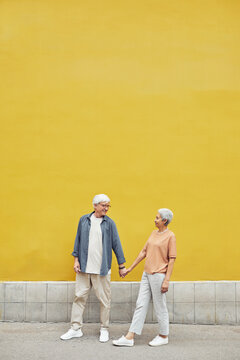 Pop color full length portrait of modern senior couple holding hands outdoors while walking against yellow background, copy space
