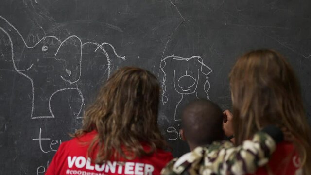Caucasian white volunteers teaching and drawing animals (Simba, lion) on the blackboard with an African child in arms. NGO Education volunteering. Africa 4K.