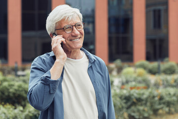Waist up portrait of modern senior man speaking by smartphone outdoors and smiling in urban city setting, copy space