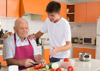 Cheerful teenage boy cooking healthy dinner together with his elderly grandfather in comfy home kitchen